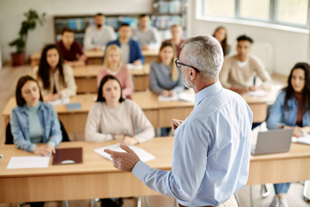 Rear view of mature teacher talking to his student during lecture at university classroom. Back view of mature professor giving lecture to large group of college students in the classroom. students without uniform at classroom stock pictures, royalty-free photos & images